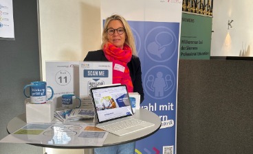 Informationsstand mit #parentsonboard-Materialen auf einem Tisch und einem Roll-up im Hintergrund. Daneben steht Katrin Peter