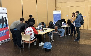 Schüler sitzen um zwei Tische und füllen ein Blatt aus, Lehrer und Coachin beobachten von der Seite